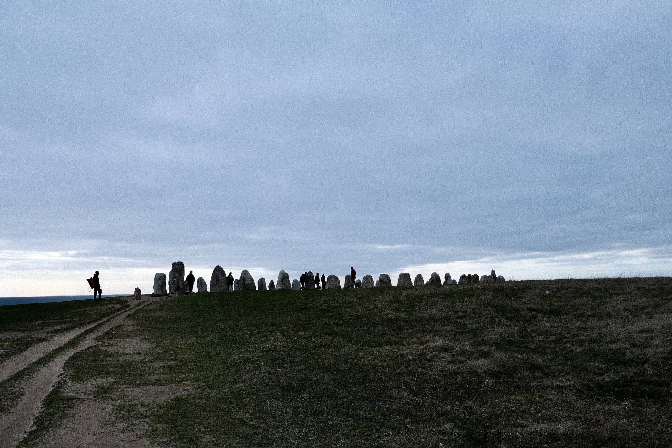 A distant view of Ales Stenar in Kåseberga, Sweden, with standing stones silhouetted along the horizon under a wide grey sky and visitors walking nearby.
