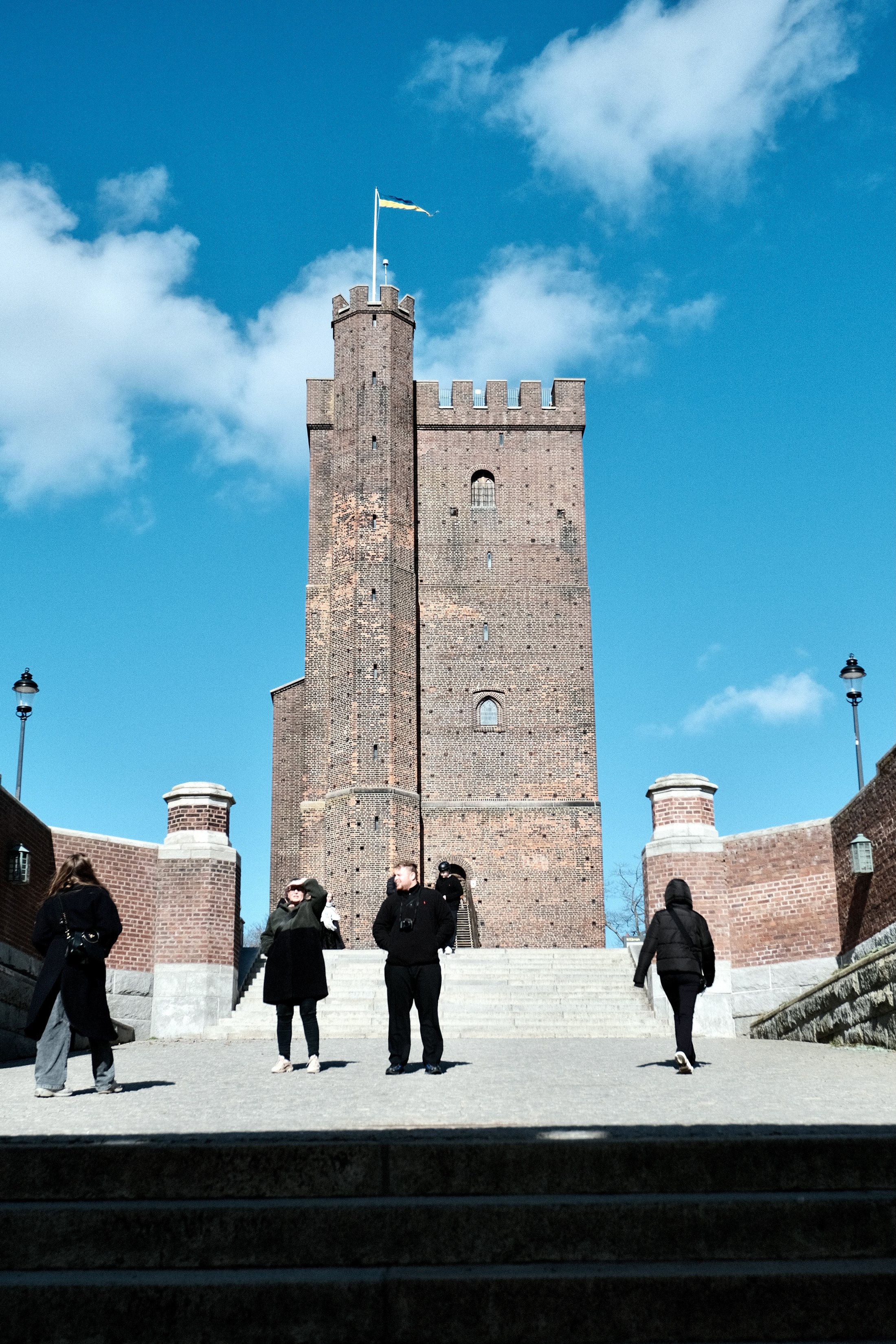 Visitors stand and climb the wide stone terrace steps leading up to the tall brick keep of Kärnan in Helsingborg, which flies a blue-and-yellow flag against a bright blue spring sky.