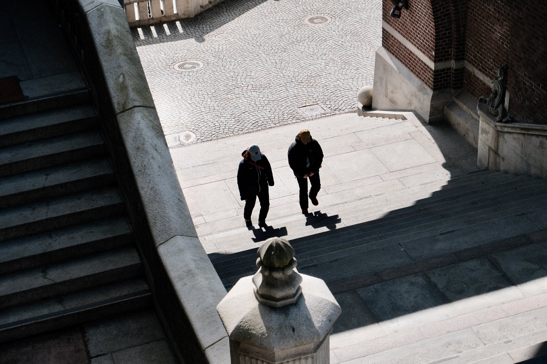 Seen from above, two people climb a broad stone staircase toward Kärnan in Helsingborg, their long shadows falling across the steps beside a fan-patterned cobblestone landing in bright spring light.
