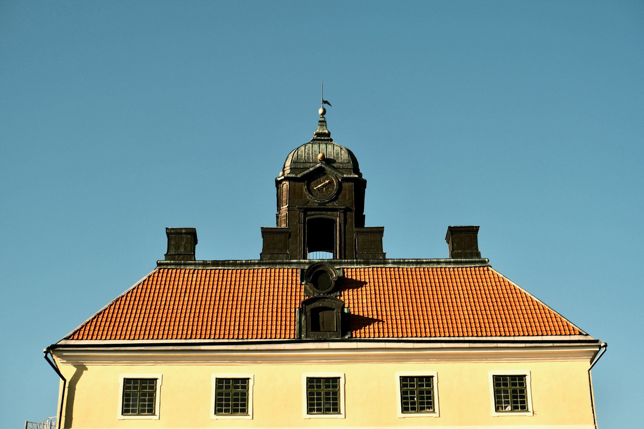 The upper storey of Engsö Castle at Ängsö, Västerås, Sweden, with its orange tile roof, four chimneys, round oculus window, and a dark clock tower and cupola against a clear sky.