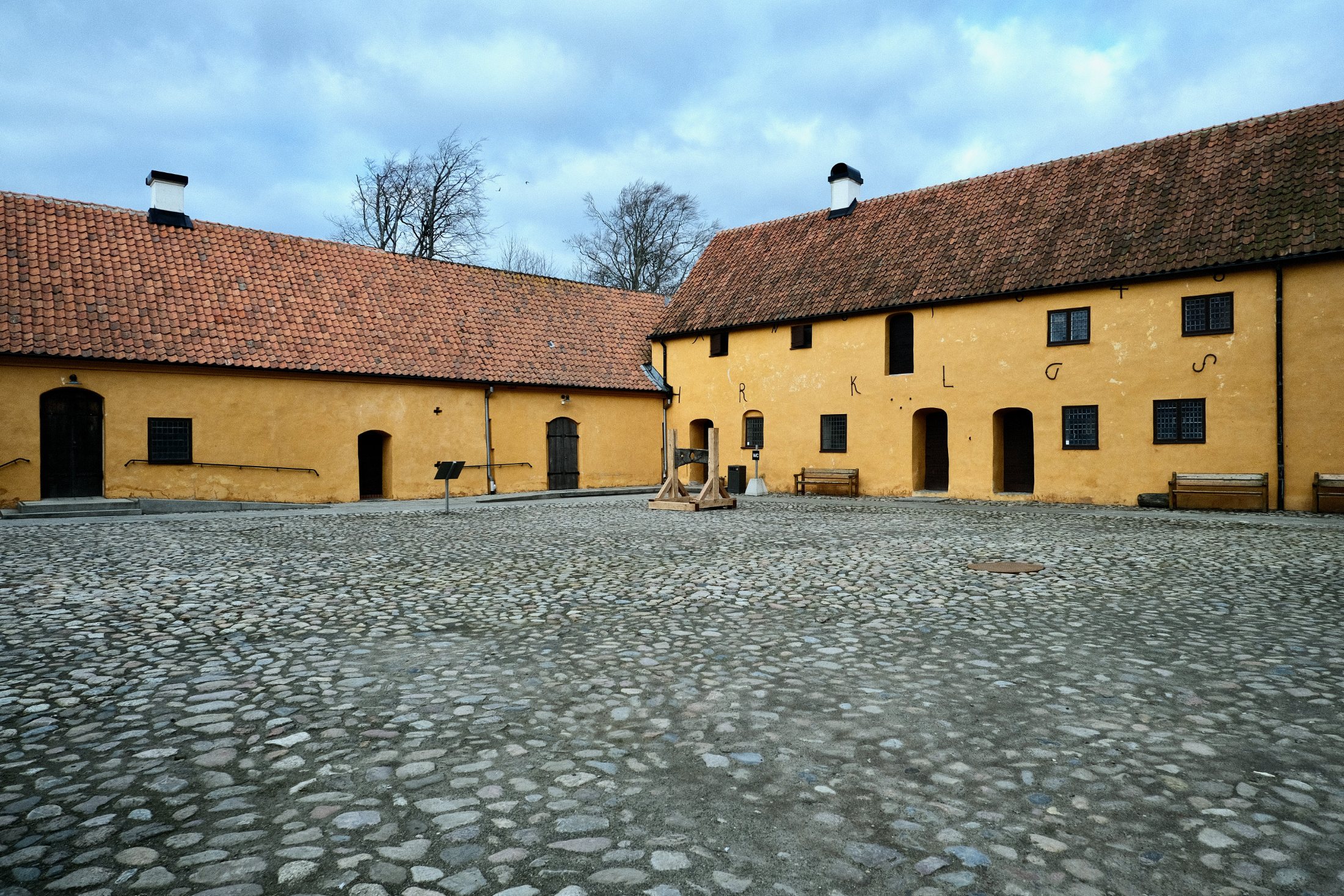 The empty cobblestone courtyard of Glimmingehus, enclosed by ochre-yellow outbuildings with red tile roofs, with a wooden replica pillory standing at its centre under an overcast spring sky.