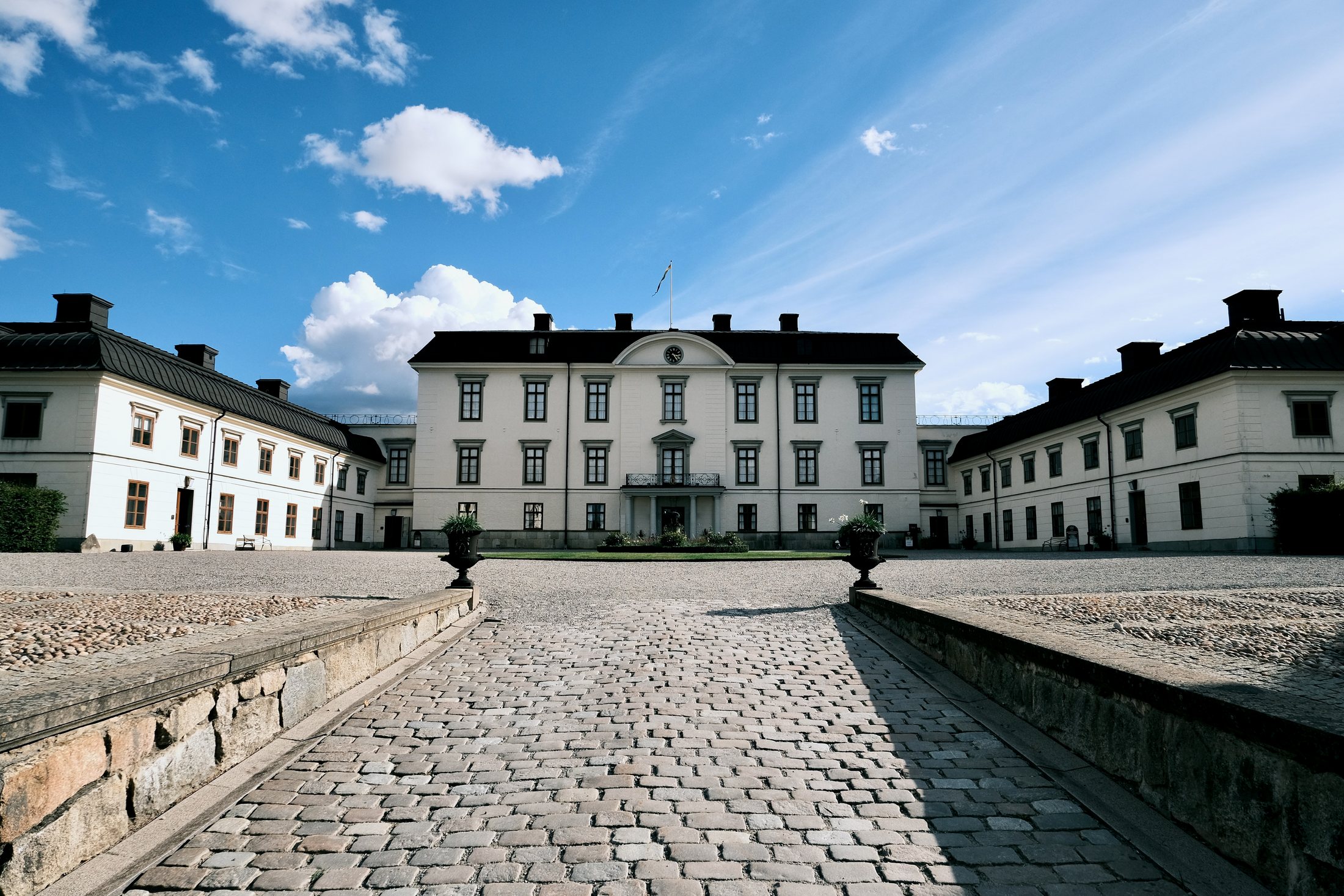 A cobbled path running into the courtyard of Rosersbergs Slott between two stone urns, with the pale main block and its clock pediment ahead and low wings closing the square on both sides.