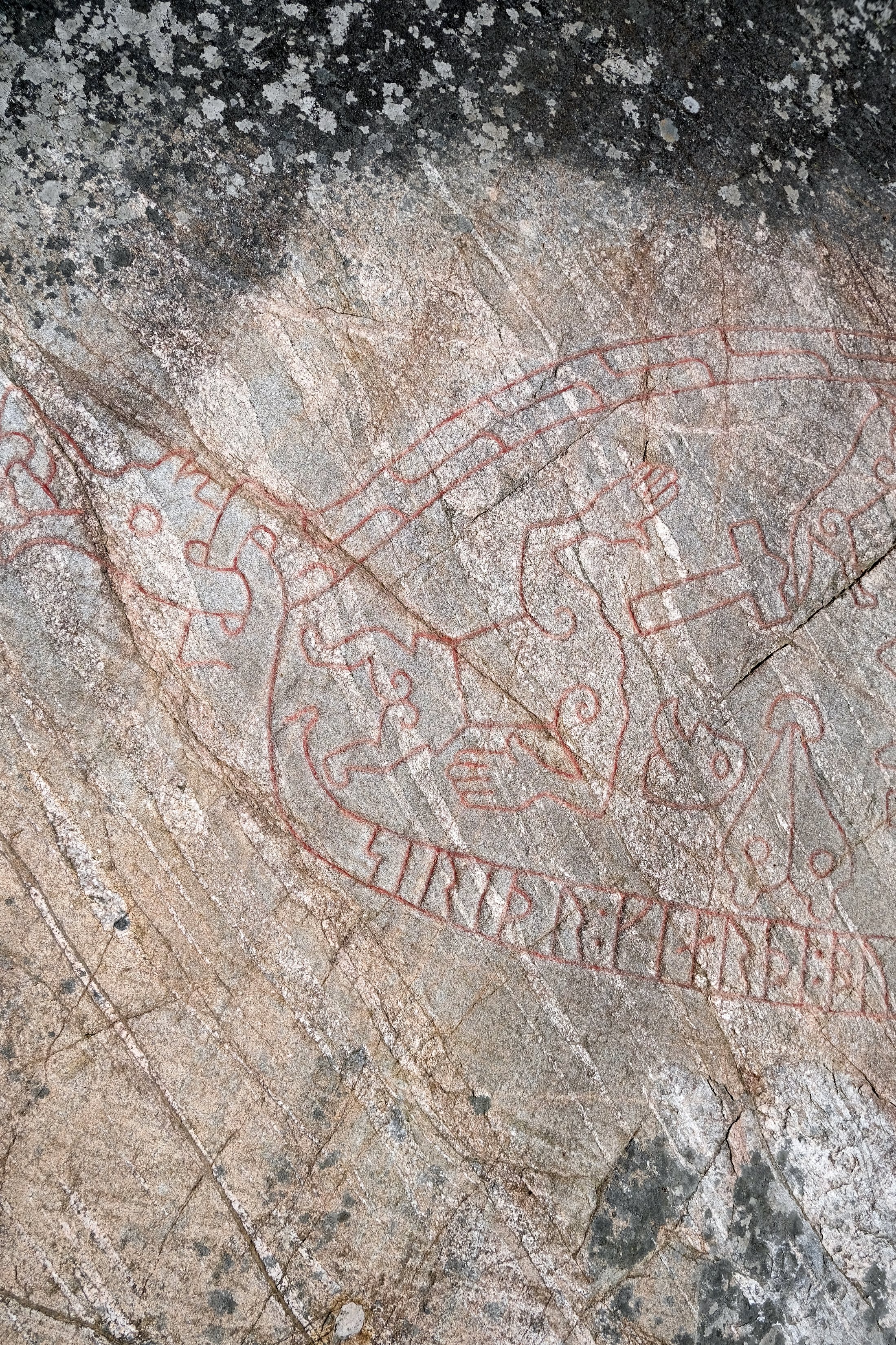 A close detail of the Sigurd Carving: red-painted runes run along a curving band — the dragon's body — beside looping carved figures and a human hand, incised into weathered grey rock flecked with lichen.