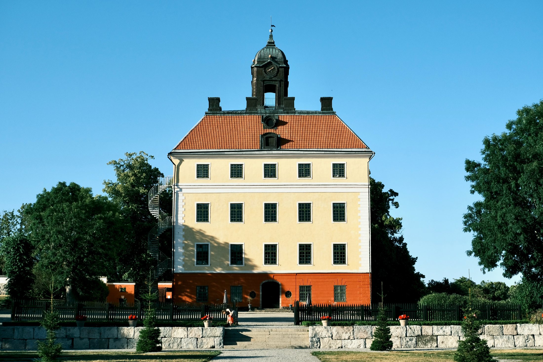 A yellow rendered castle with an orange-red base, tile roof and central clock tower at Ängsö, Västerås, Sweden, above a stone terrace and black picket fence.