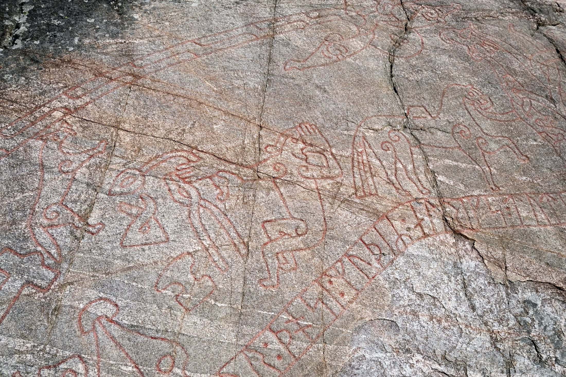 A close detail of the Sigurd Carving showing red-painted figures — interlacing beasts and a bird — above a band of runes, all incised into cracked, lichen-flecked grey rock.
