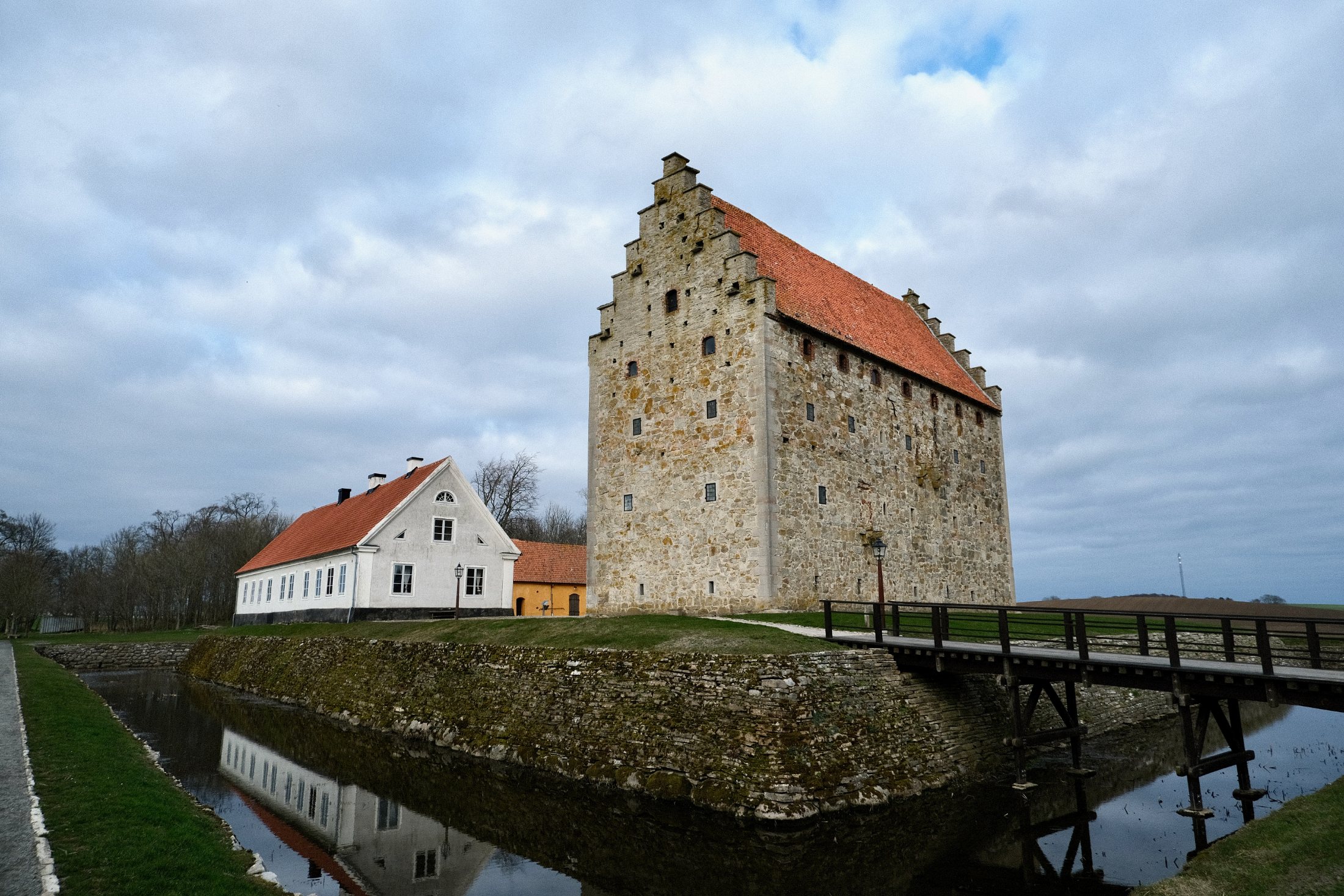The tall stone keep of Glimmingehus rises beside a white-walled manor house, reflected in the surrounding moat under an overcast spring sky, with a wooden footbridge crossing the water to the right.