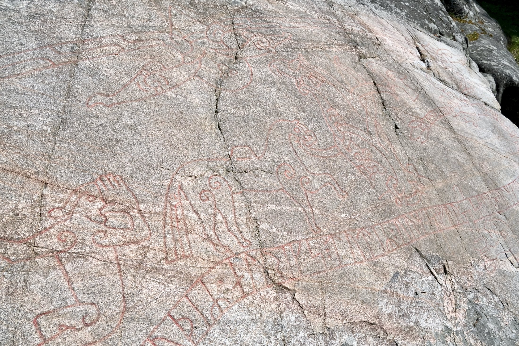A close detail of the Sigurd Carving showing red-painted figures — a laden horse and birds among looping lines — set above a long band of runes incised into the sloping grey rock.