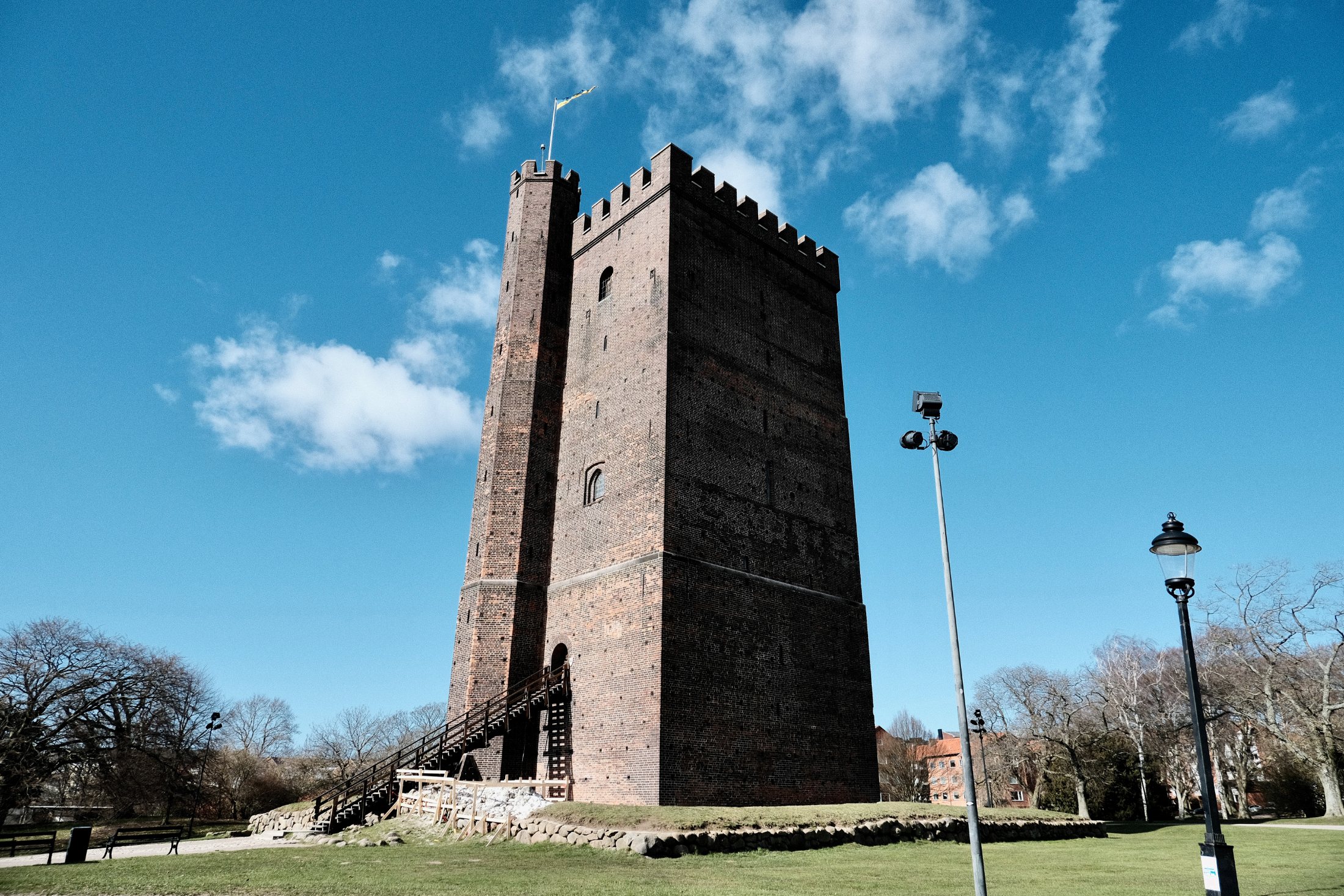 The tall square brick keep of Kärnan rises from a grassy mound in Helsingborg, its crenellated top flying a Swedish flag against a bright blue spring sky, with a wooden stair climbing to its arched doorway.