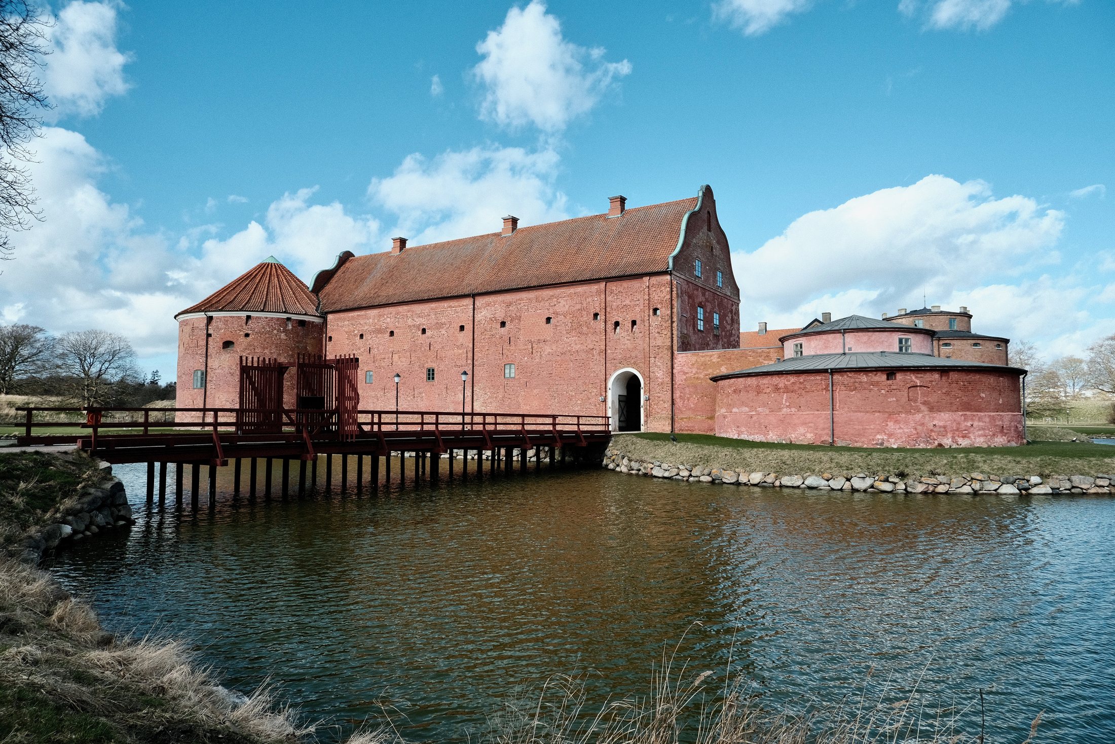 A wide view of Landskrona Slott in Sweden, with red brick castle buildings, a wooden bridge, the surrounding moat, and a bright cloudy sky.