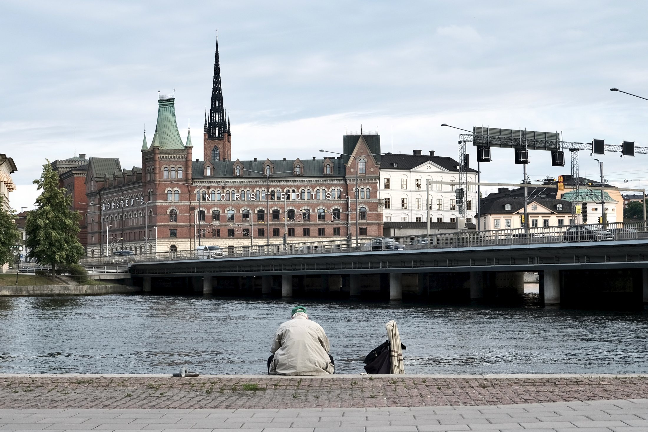A view from Klara Mälarstrand in Stockholm looking across the water toward Riddarholmen, with an elderly man sitting alone by the waterfront in the foreground.