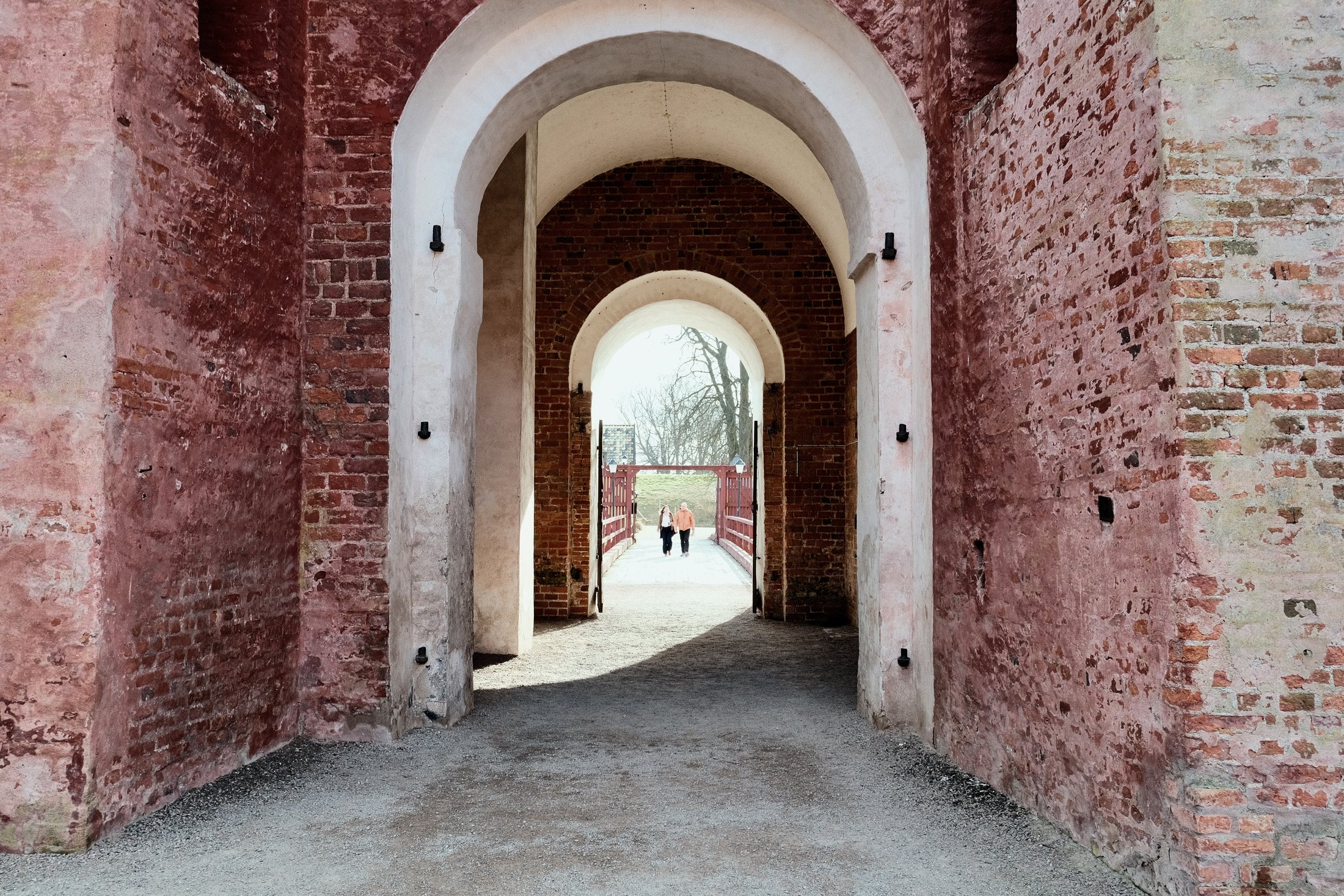 An arched passage inside Landskrona Slott, with red brick walls, white archways, and two people walking across the bridge in the distance.
