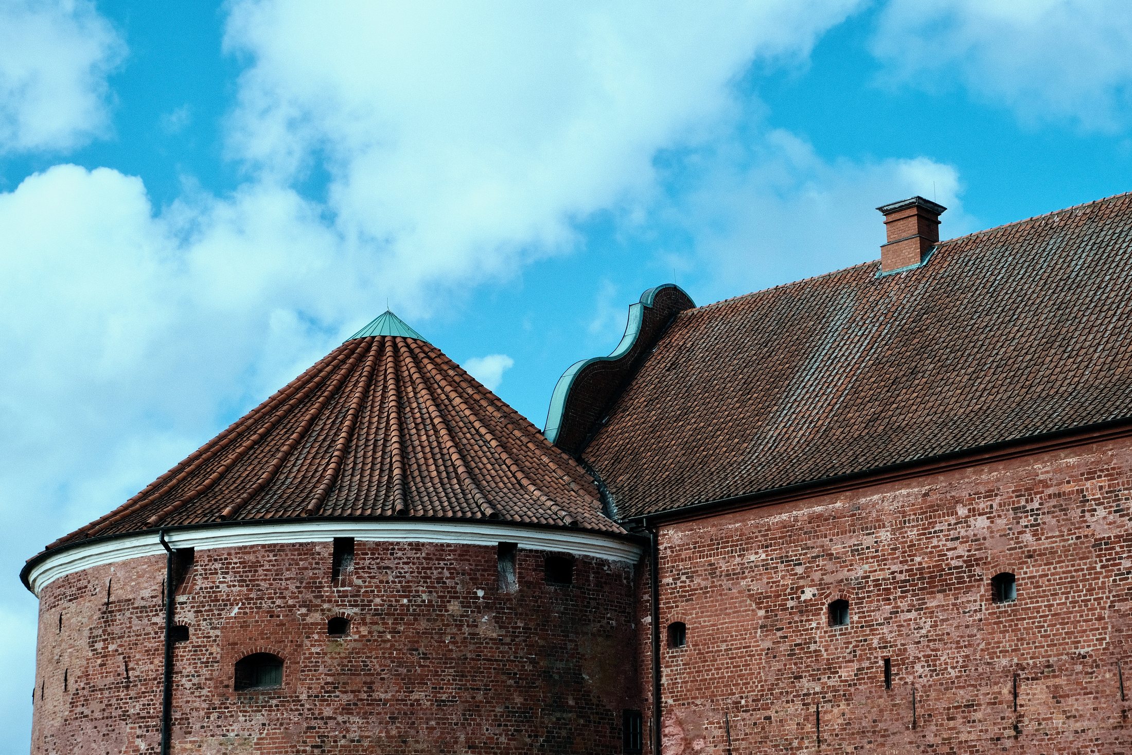 A close architectural view of Landskrona Slott, showing red brick walls, tiled rooflines, a round tower roof, and blue sky with clouds above.