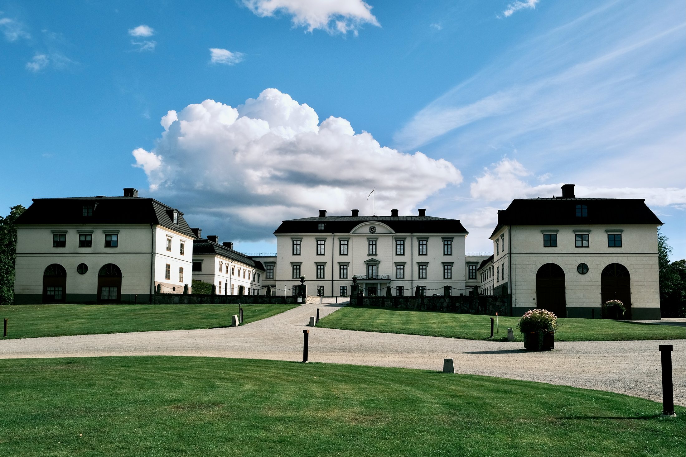 The pale façades and black roofs of Rosersbergs Slott across its gravel forecourt, the main block carrying a clock in its pediment between two wing pavilions with arched carriage doors.