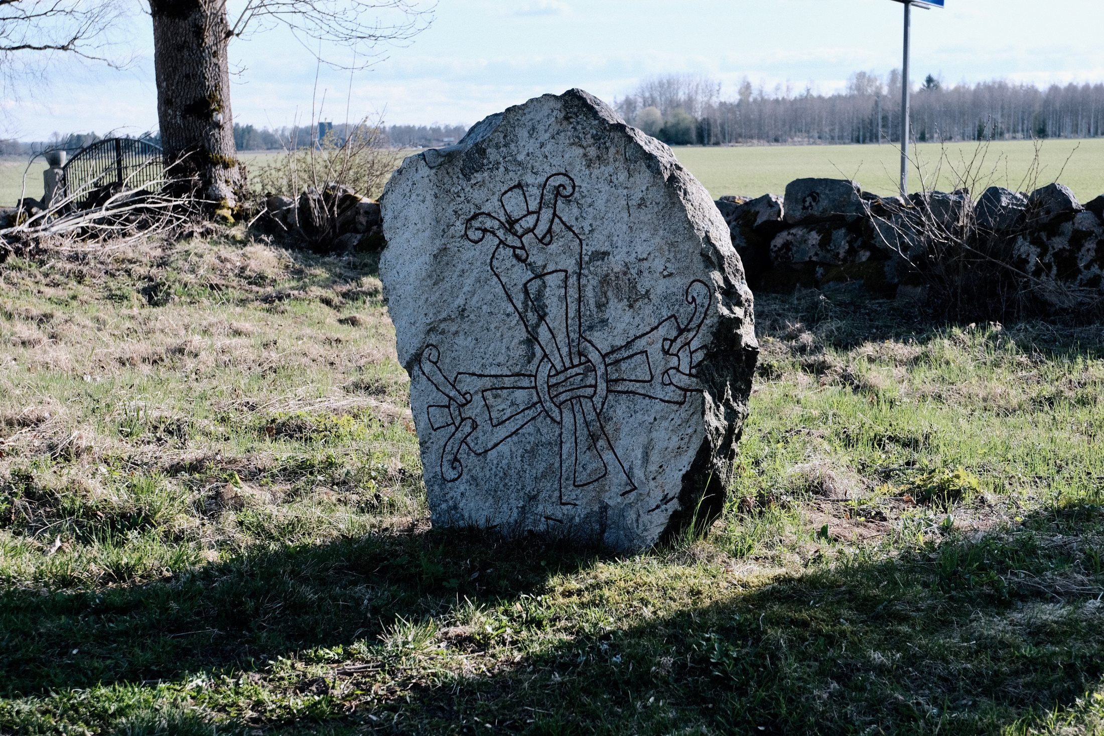 A weathered gray rune stone carved with an interlaced runic cross motif, standing in a grass field near a stone wall and bare tree in Rytterne, Västerås, Sweden.