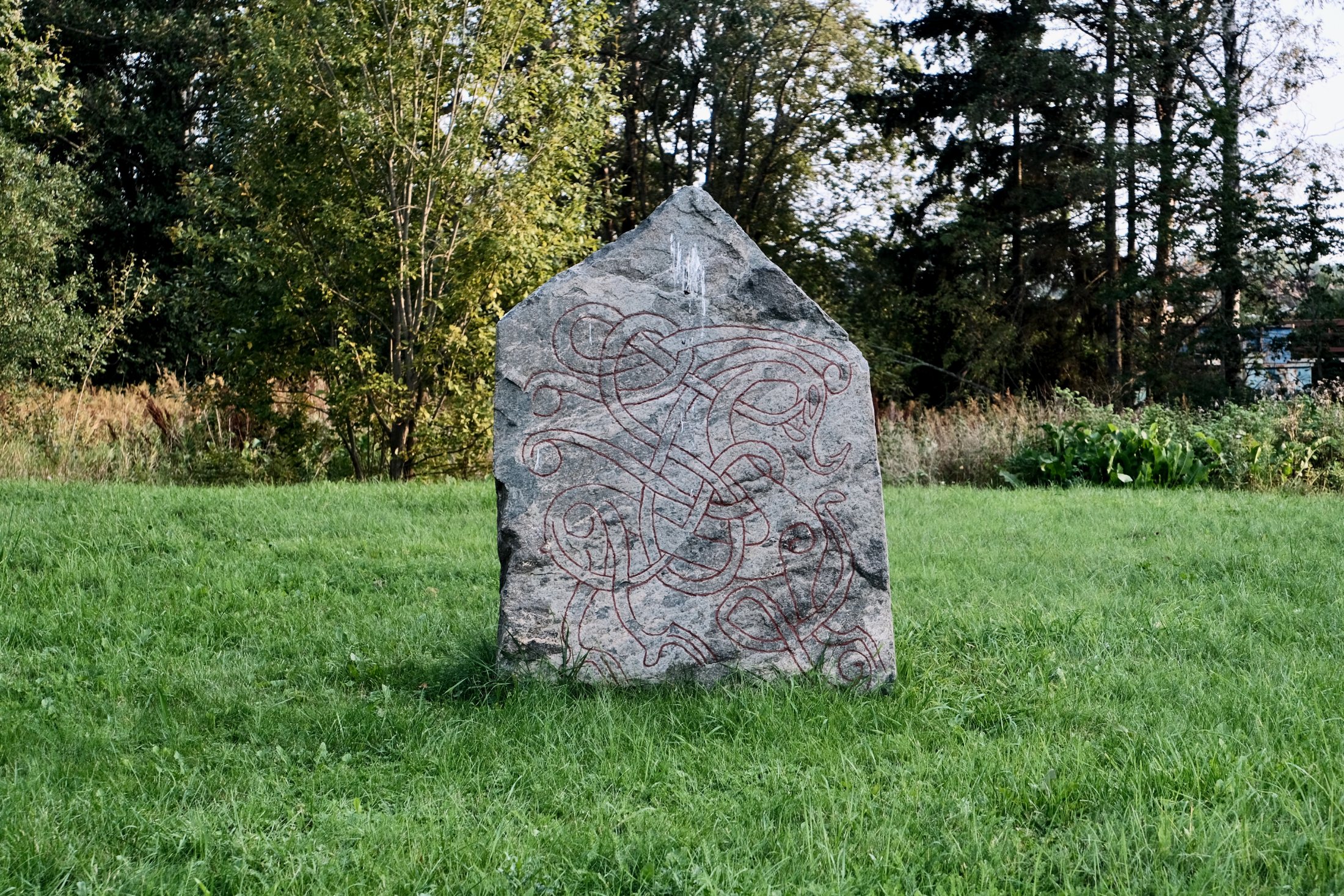 A gabled grey granite runestone stands in a green late-summer meadow, its face covered by an interlaced serpent design painted deep red, with leafy trees behind it.