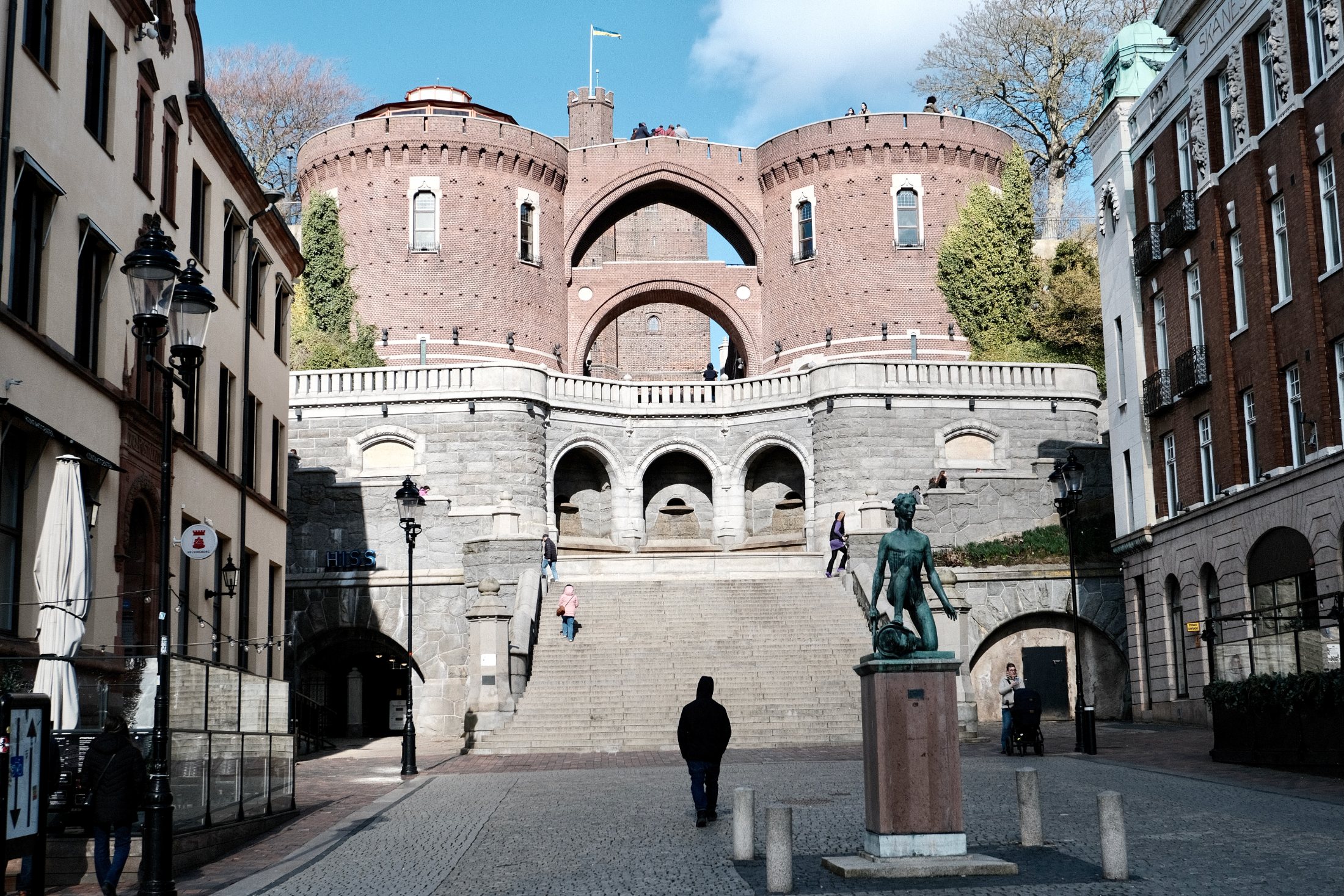 A monumental brick-and-stone terrace staircase rises from a cobbled Helsingborg square toward the twin round towers and the Kärnan keep above, with a bronze statue and a few people climbing the steps in bright spring light.