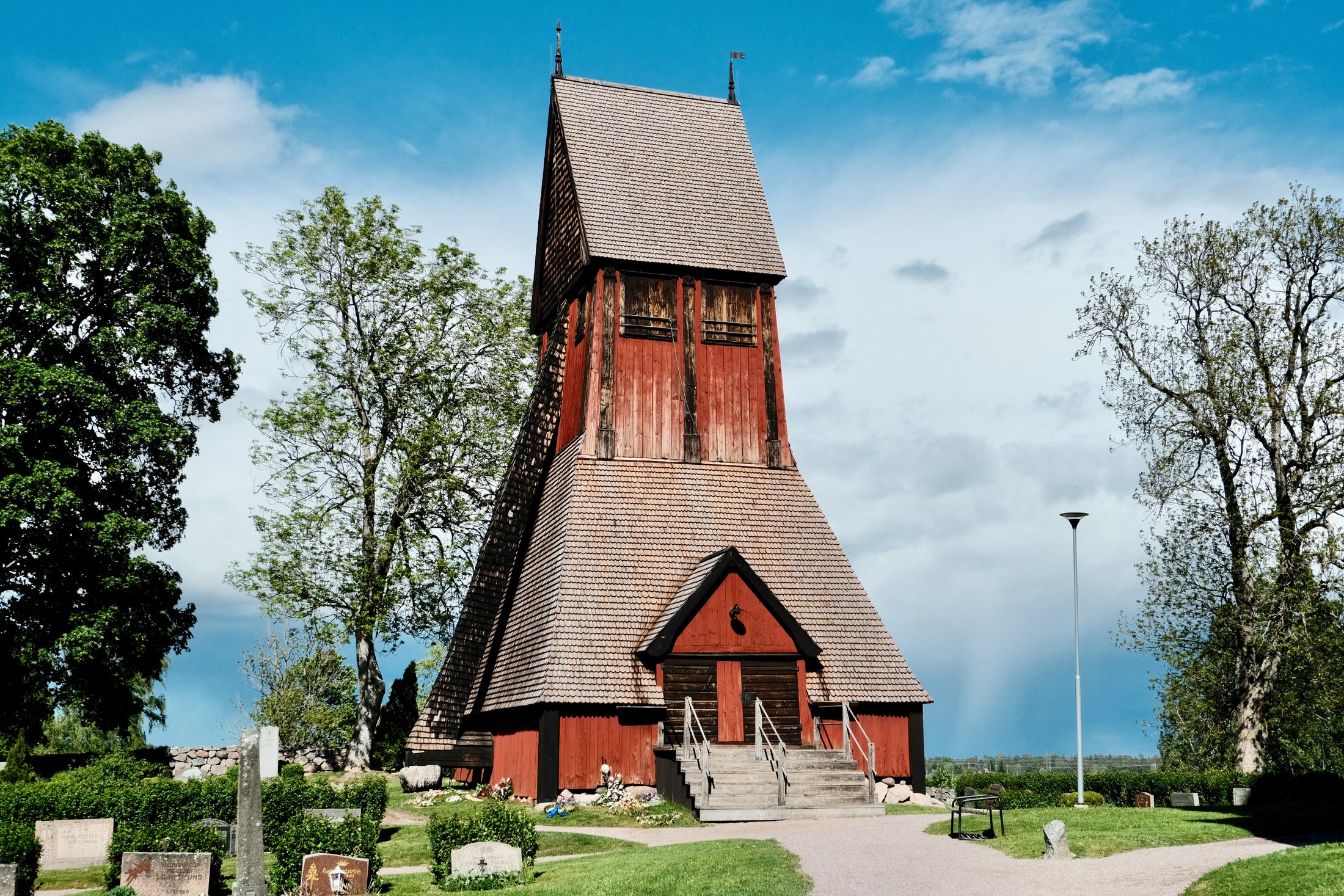 The wooden bell tower beside the Church of Old Uppsala in Sweden, standing among trees, gravestones, grass, and a bright spring sky.