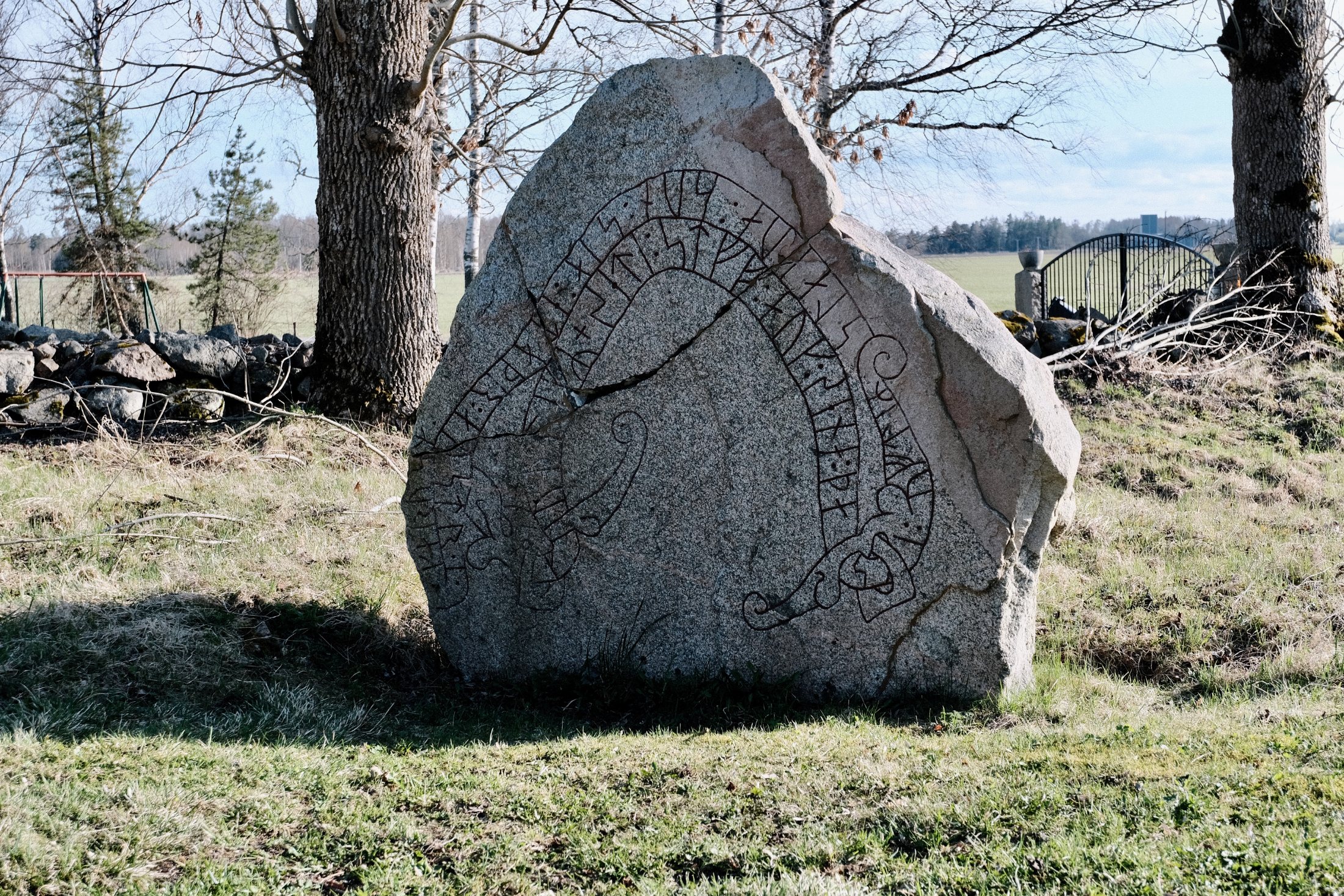 A granite rune stone carved with a curved runic inscription band, standing beside a large tree with a stone wall, iron gate, and open field behind it in Rytterne, Västerås, Sweden.