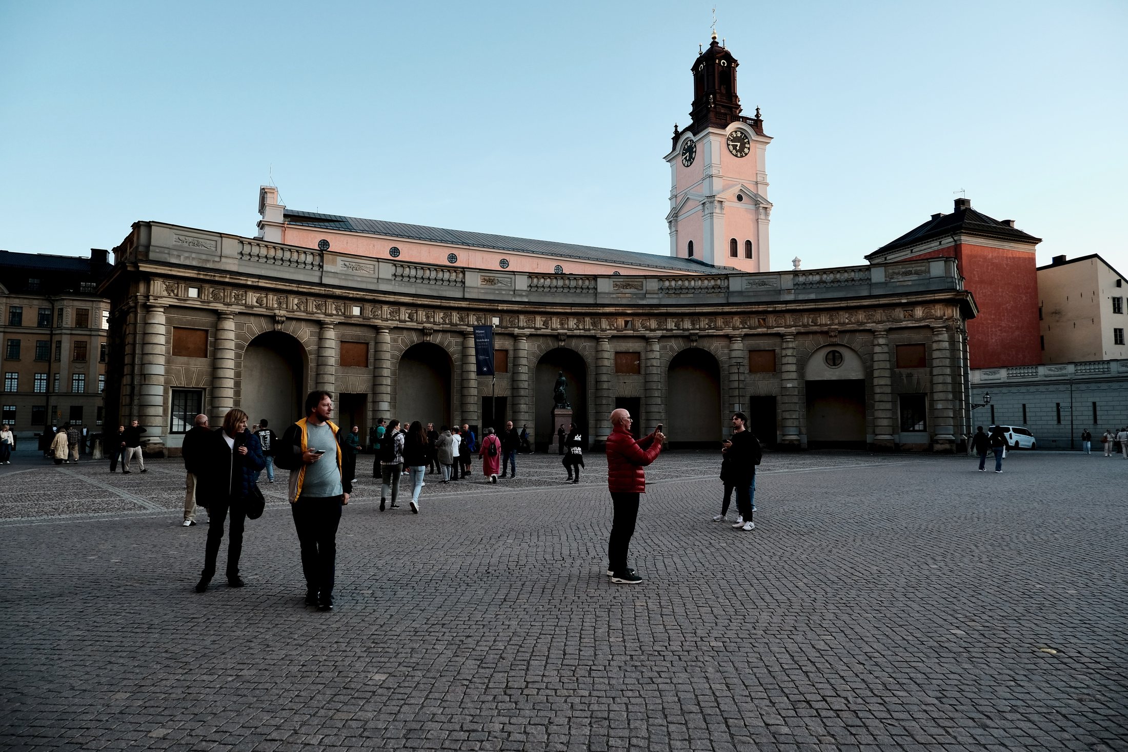 A view from Yttre Borggården in Gamla Stan, Stockholm, with people standing in the courtyard and the tower of Stockholm Cathedral visible beyond the palace buildings.
