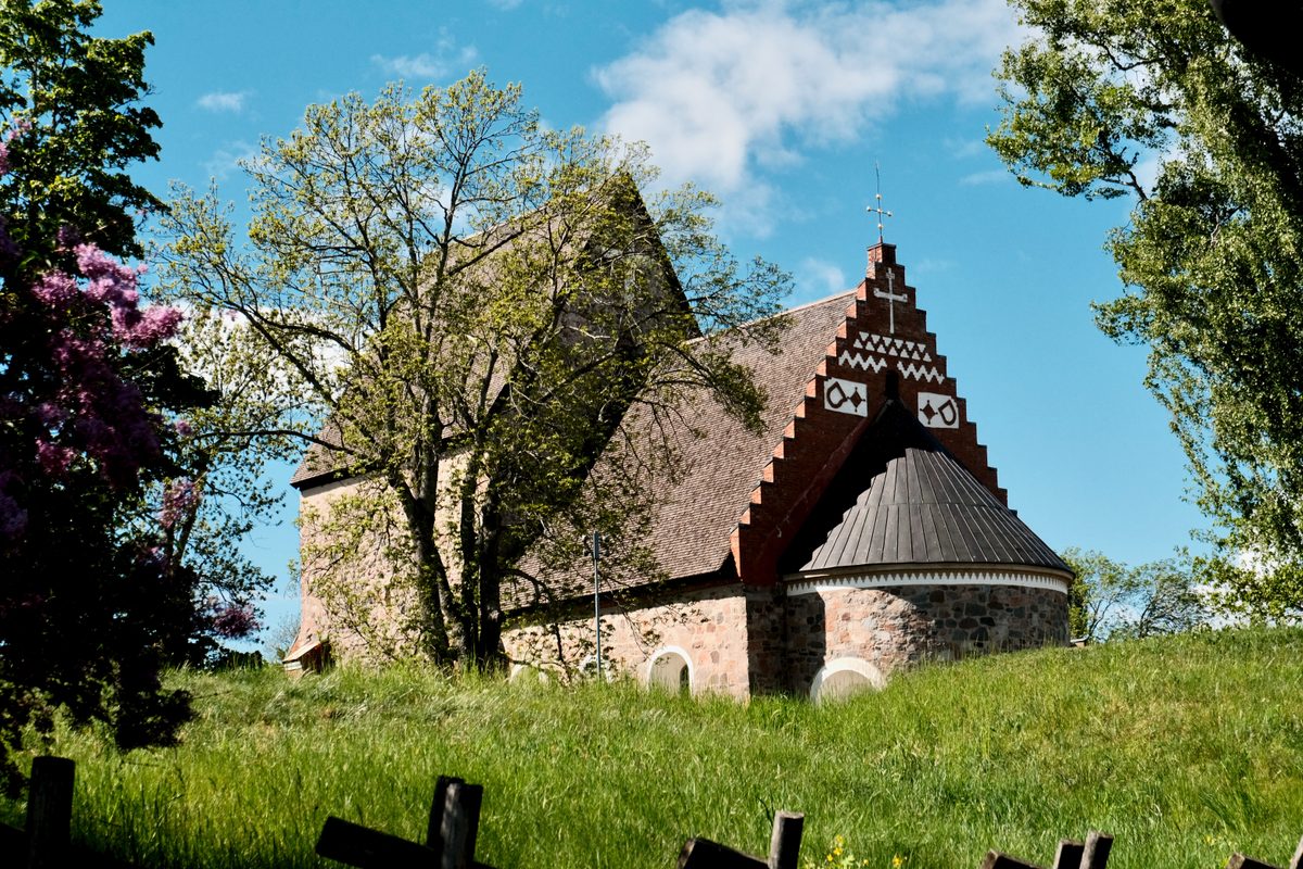 The Church of Old Uppsala in Sweden, seen on a grassy hill surrounded by trees, with stone walls, a red brick gable, and a blue sky above.