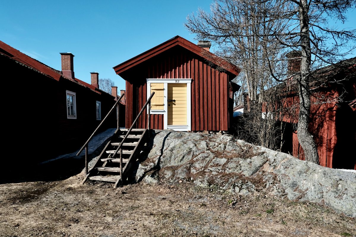 A red wooden cottage at Lövånger Kyrkstad in Västerbotten, Sweden, standing on rocky ground with a yellow door, wooden steps, bare trees, and a clear blue sky.