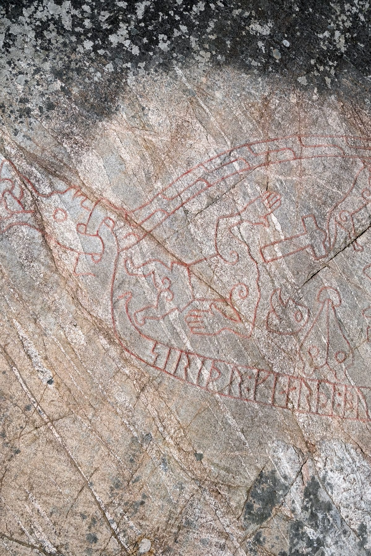 A close detail of the Sigurd Carving: red-painted runes run along a curving band — the dragon's body — beside looping carved figures and a human hand, incised into weathered grey rock flecked with lichen.