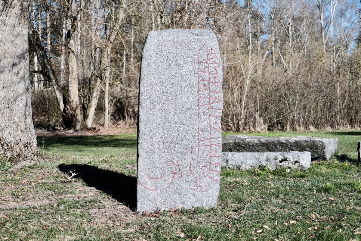 A tall gray granite rune stone with a vertical band of red-painted runes carved down its right edge, standing near a tree and low granite benches on a lawn in Dingtuna, Västerås, Sweden.