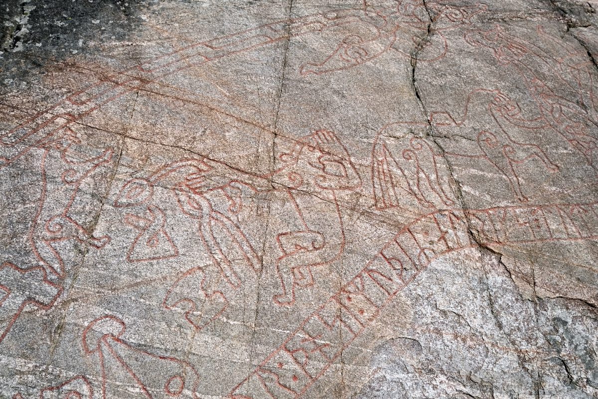 A close detail of the Sigurd Carving showing red-painted figures — interlacing beasts and a bird — above a band of runes, all incised into cracked, lichen-flecked grey rock.