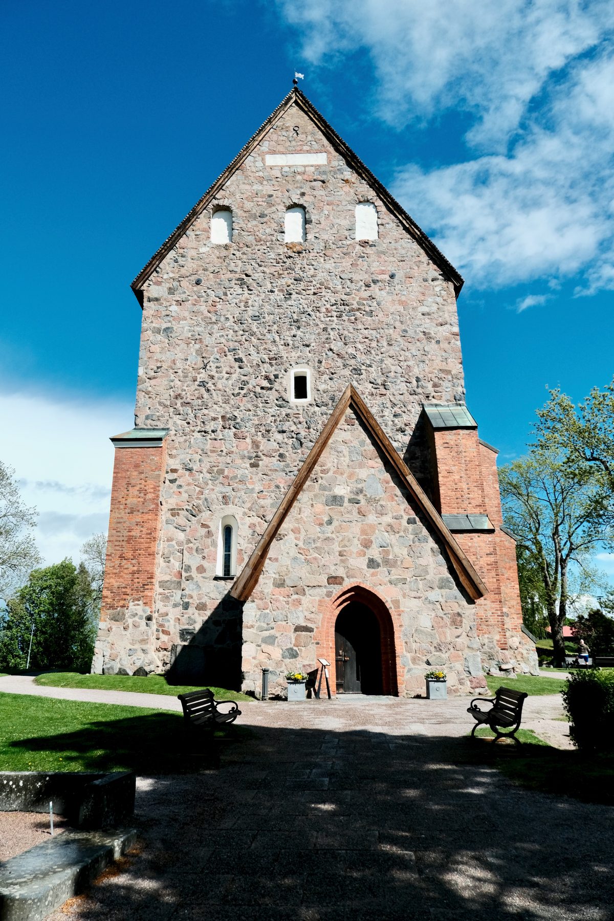 The front façade of the Church of Old Uppsala in Sweden, showing a tall medieval stone church with a red brick entrance, benches, green grass, and a blue sky with clouds.