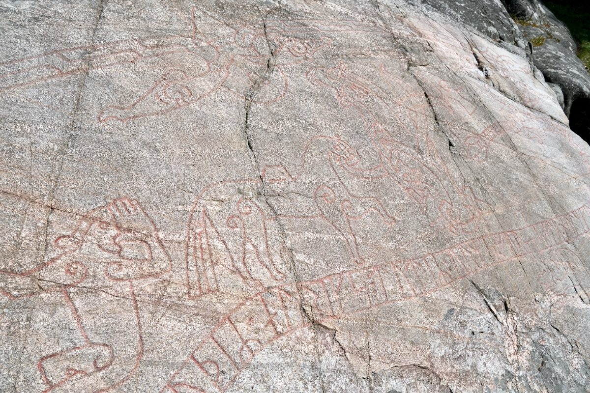 A close detail of the Sigurd Carving showing red-painted figures — a laden horse and birds among looping lines — set above a long band of runes incised into the sloping grey rock.