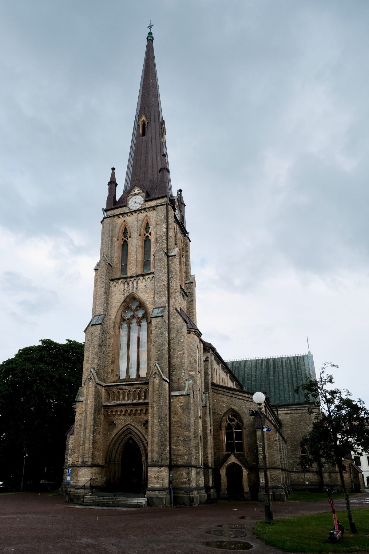 The yellow brick tower and tall spire of Haga Kyrka in Gothenburg, Sweden, seen from below against an overcast sky, with Gothic arched windows and a green copper roof.