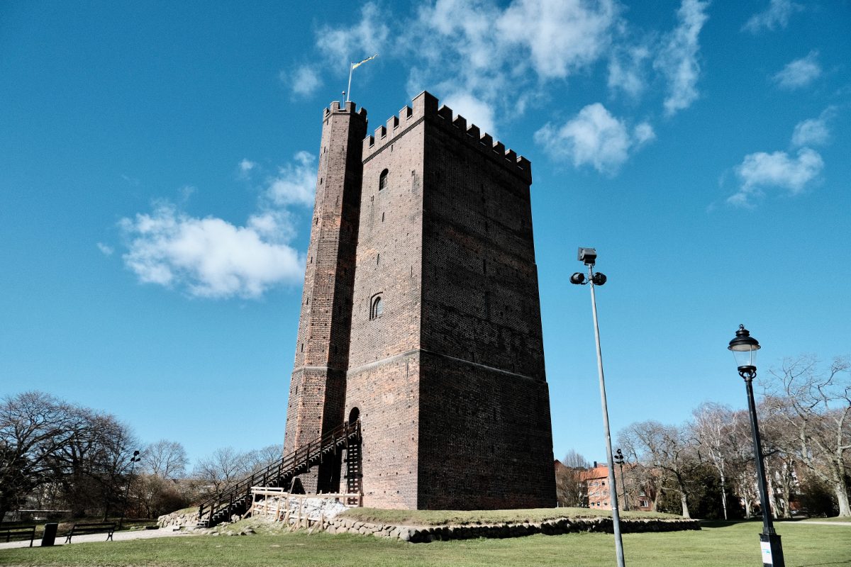 The tall square brick keep of Kärnan rises from a grassy mound in Helsingborg, its crenellated top flying a Swedish flag against a bright blue spring sky, with a wooden stair climbing to its arched doorway.