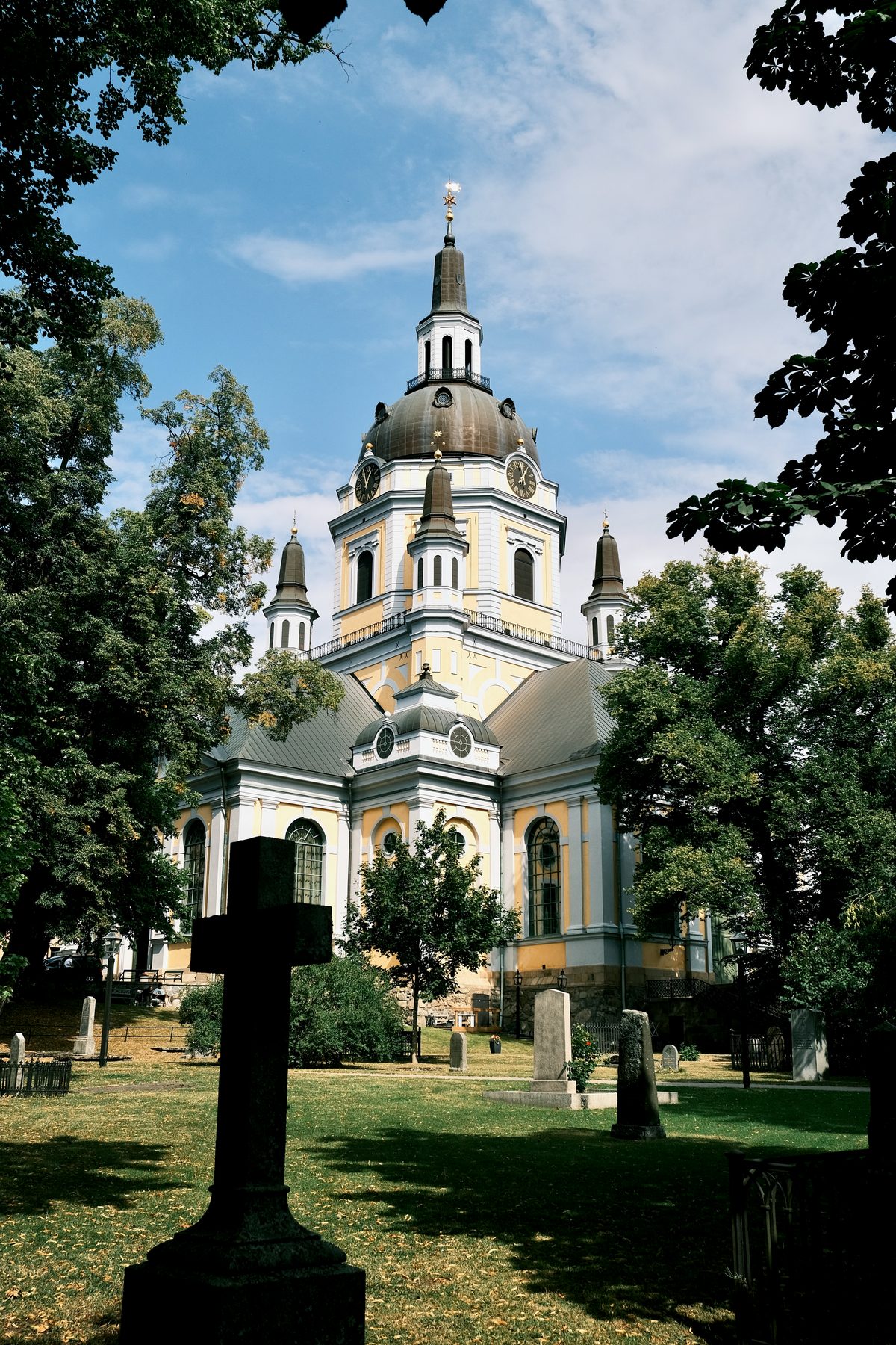 Katarina Kyrka in Södermalm, Stockholm, seen from its churchyard through summer foliage, with a copper dome, clock tower, yellow and white façade, and stone gravestones on the grass.