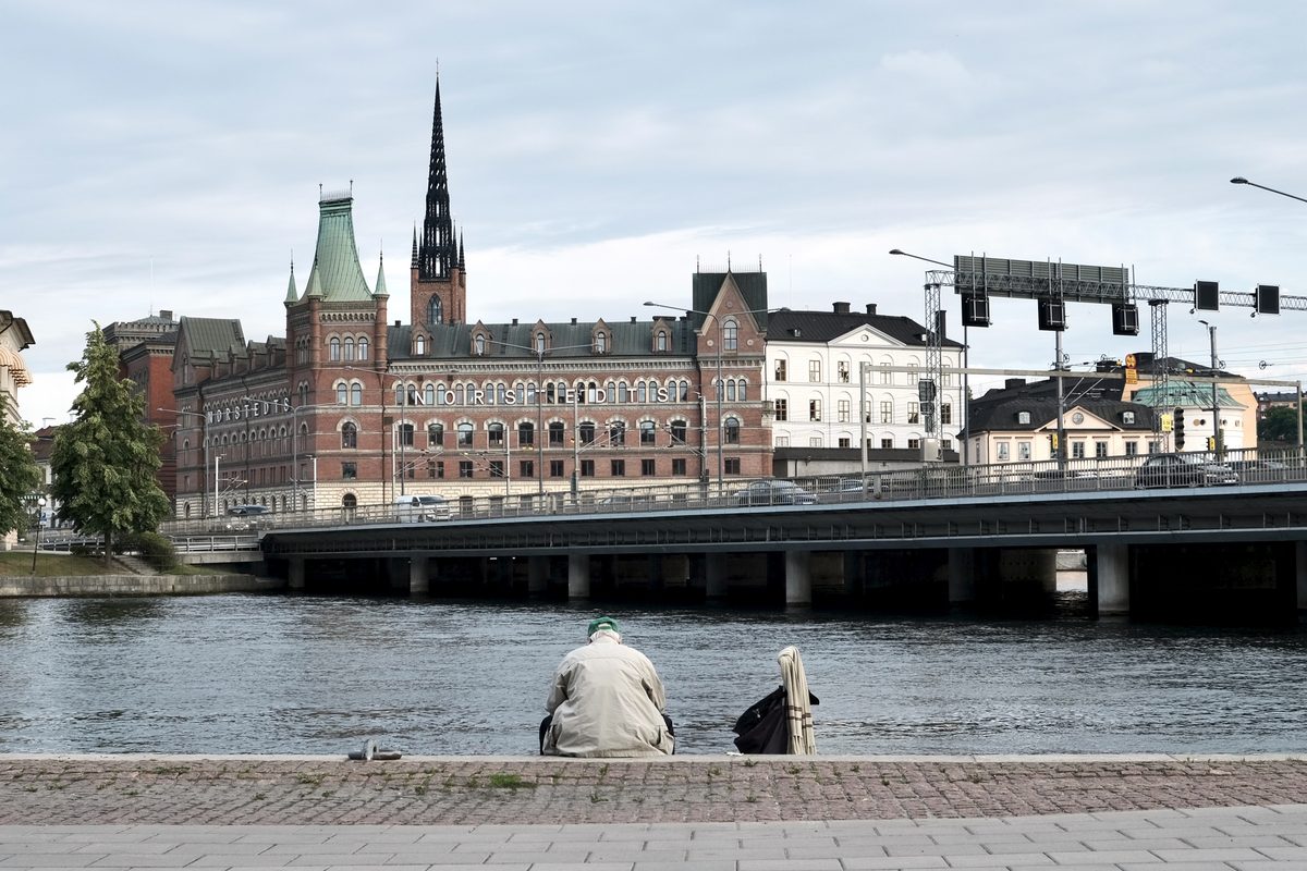 A view from Klara Mälarstrand in Stockholm looking across the water toward Riddarholmen, with an elderly man sitting alone by the waterfront in the foreground.