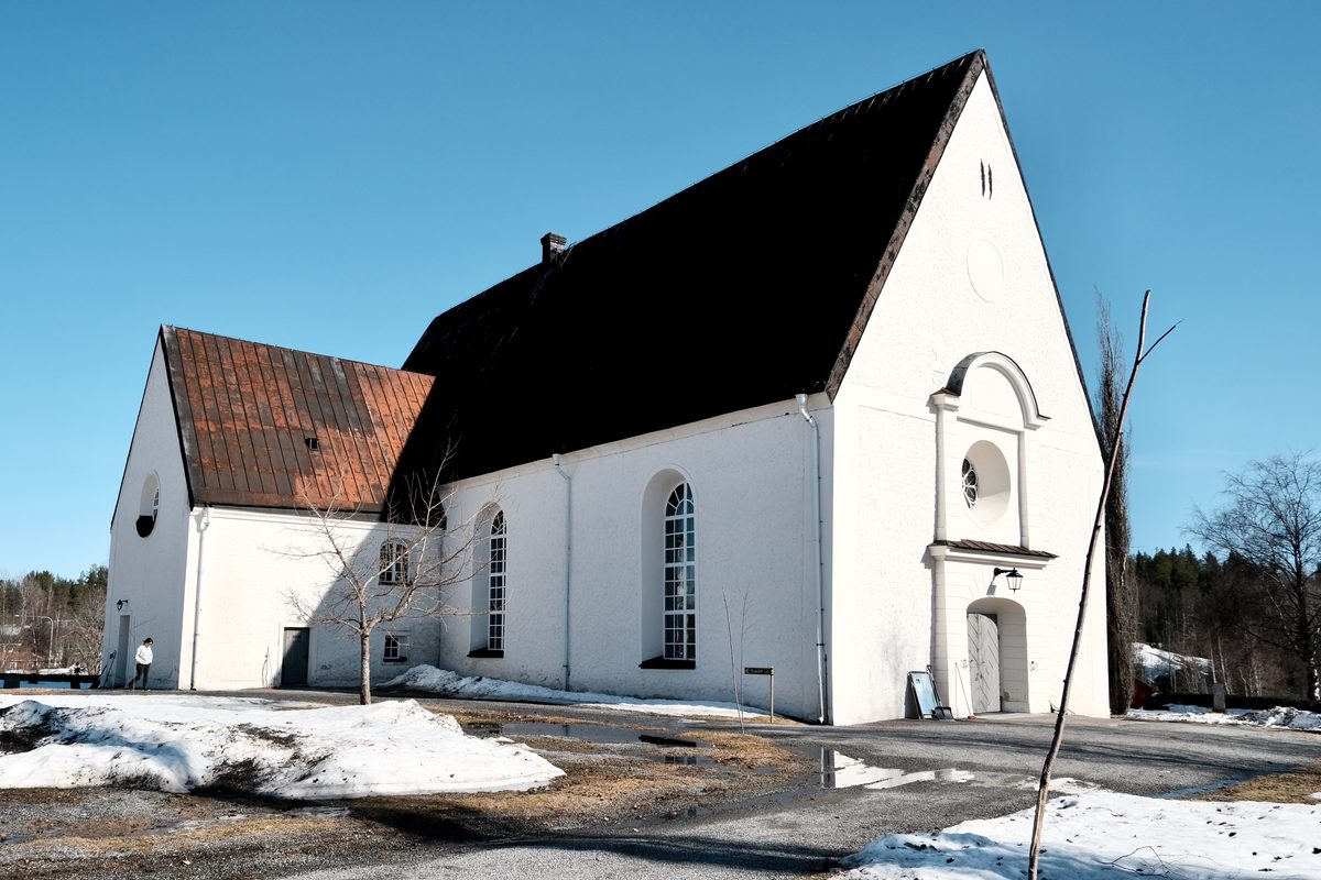 Lövånger Church in Västerbotten, Sweden, seen from the side with white walls, a dark roof, patches of snow, bare trees, and a clear blue spring sky.