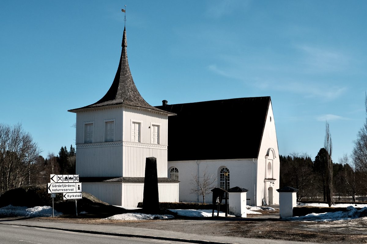 Lövånger Church in Västerbotten, Sweden, with a white church building, wooden bell tower, road signs, patches of snow, and a clear blue sky.