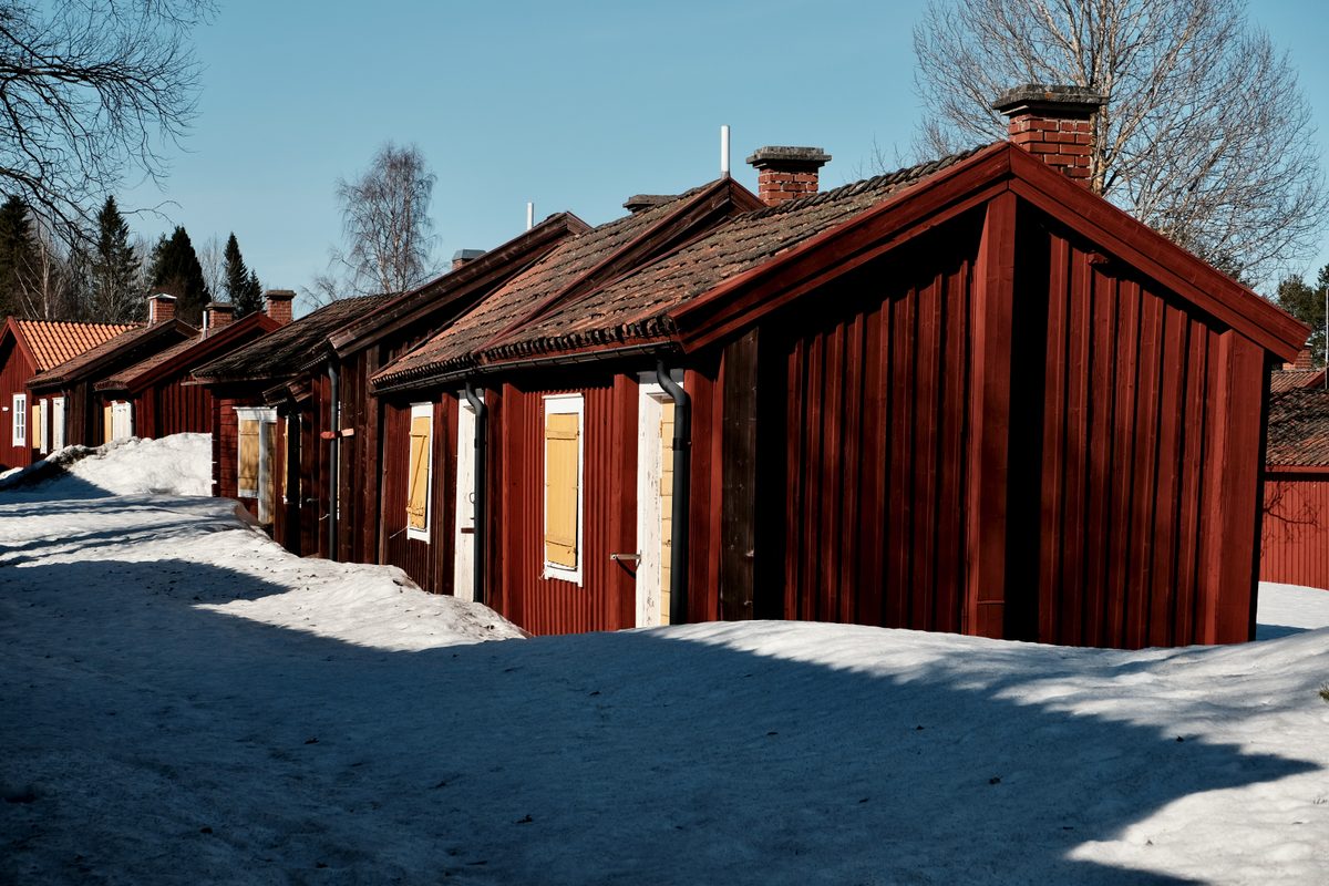 Red wooden church town cottages at Lövånger Kyrkstad in Västerbotten, Sweden, standing in spring snow under a clear blue sky.