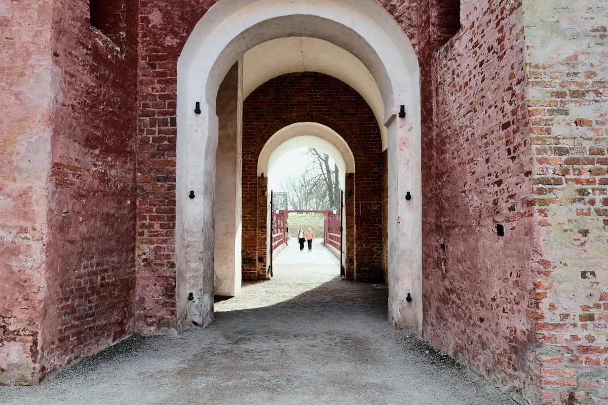 An arched passage inside Landskrona Slott, with red brick walls, white archways, and two people walking across the bridge in the distance.