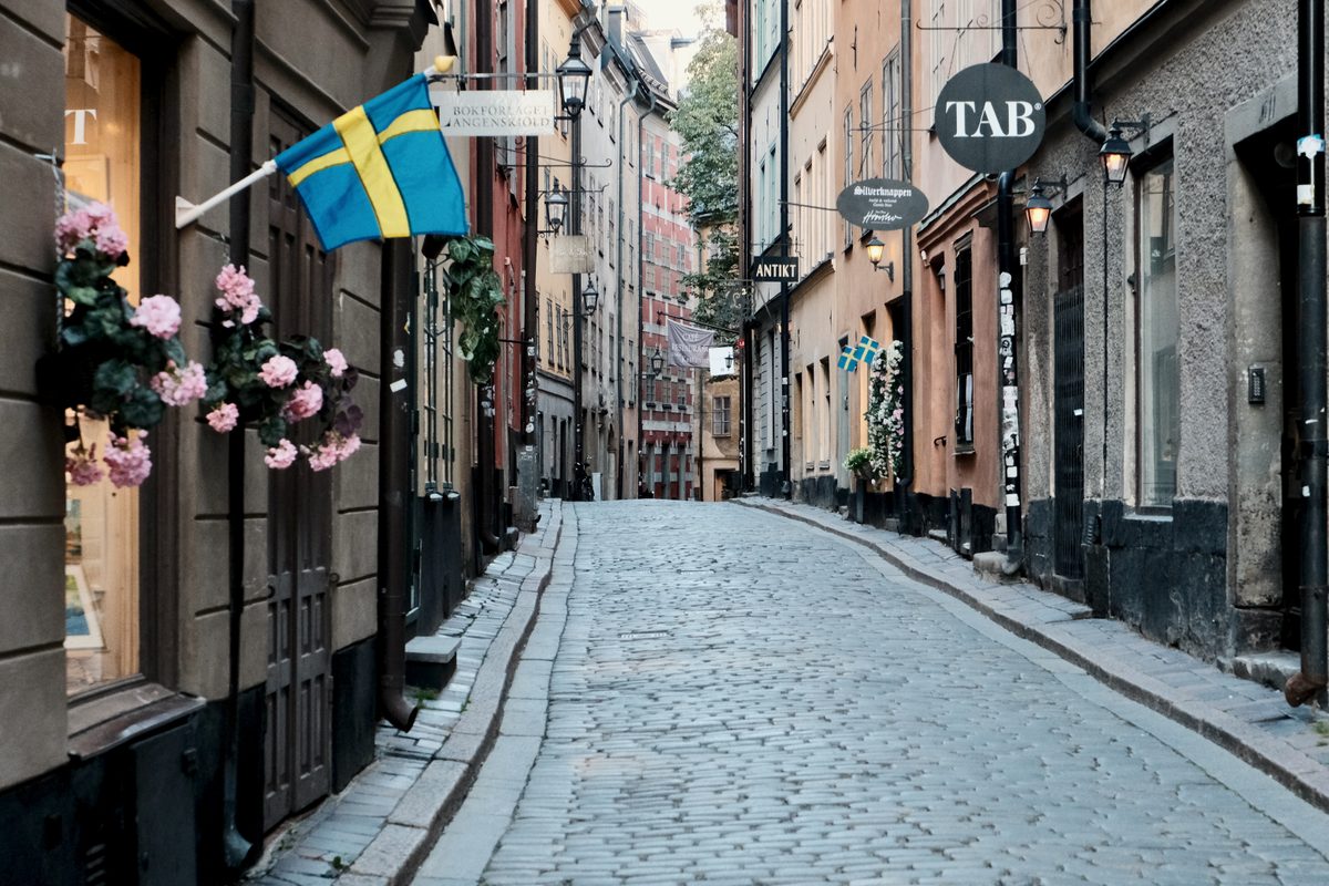 A quiet cobblestone street in Gamla Stan, Stockholm, lined with historic buildings, hanging signs, street lamps, flowers, and Swedish flags.