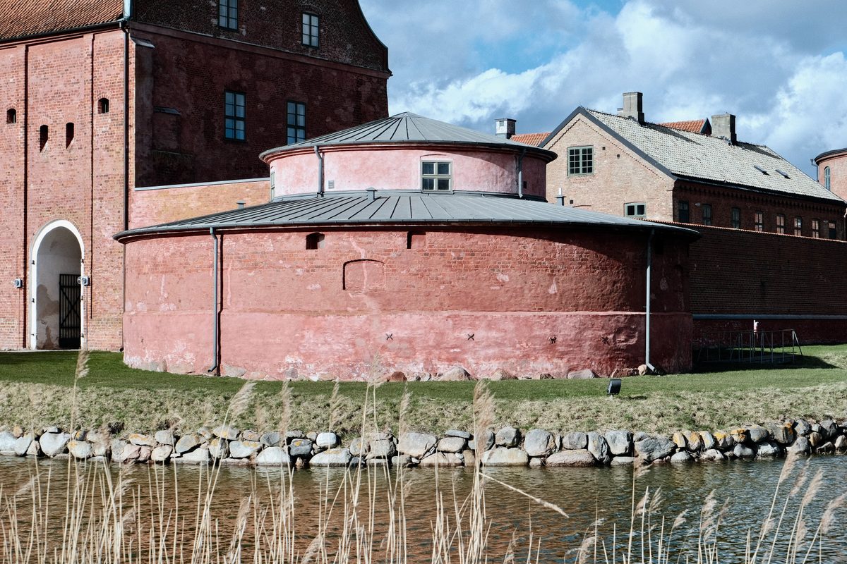 A round red brick bastion at Landskrona Slott in Sweden, seen beside the water with historic castle buildings and a cloudy sky in the background.