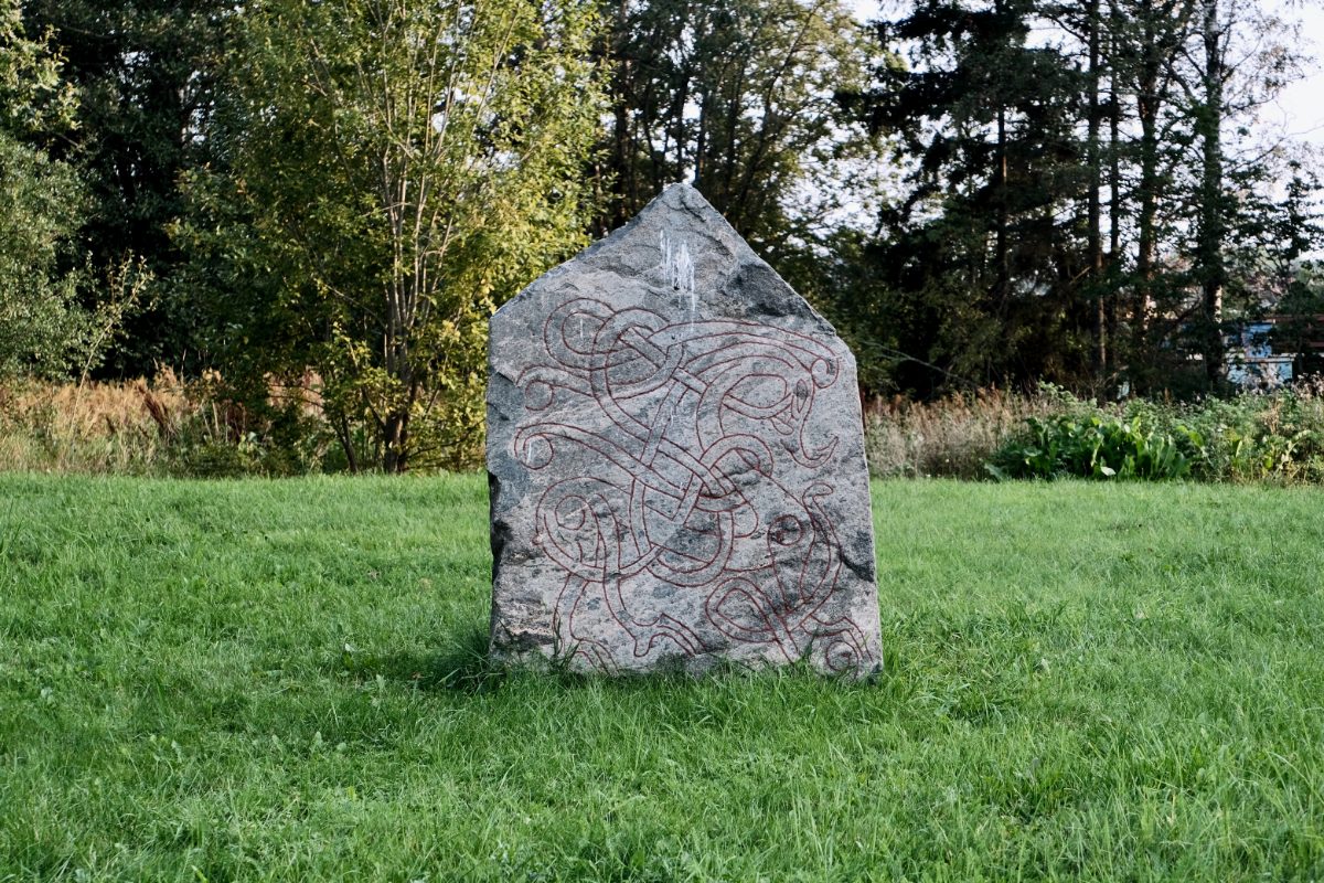 A gabled grey granite runestone stands in a green late-summer meadow, its face covered by an interlaced serpent design painted deep red, with leafy trees behind it.