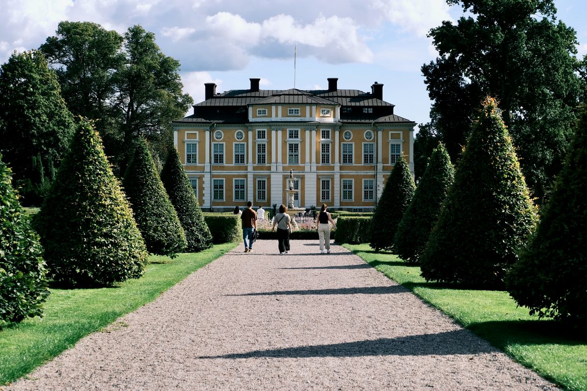 A gravel avenue in the baroque garden of Steninge Slott, lined with clipped conical trees, running past a fountain toward the ochre palace façade as a few visitors walk up the path.