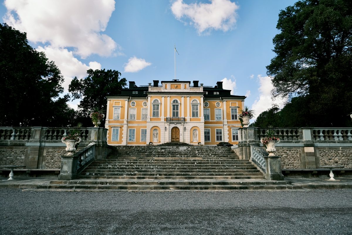 The ochre and white baroque façade of Steninge Slott seen from the gravel drive, a broad stone staircase and urn-lined balustrade climbing to the entrance under a Swedish flag.