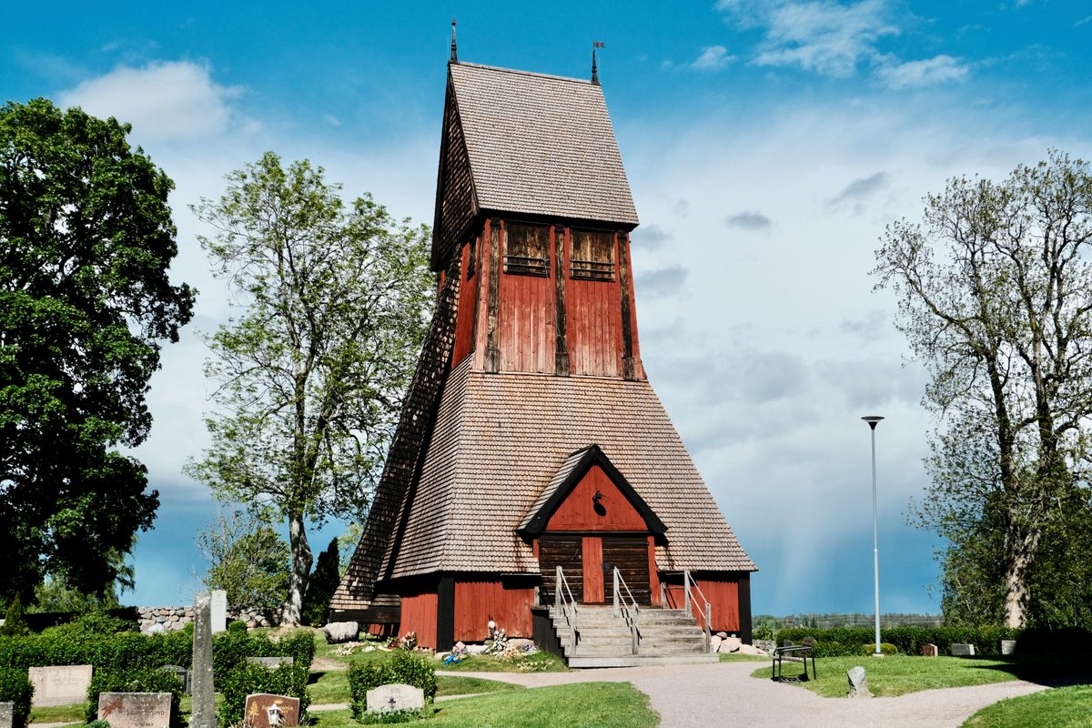 The wooden bell tower beside the Church of Old Uppsala in Sweden, standing among trees, gravestones, grass, and a bright spring sky.