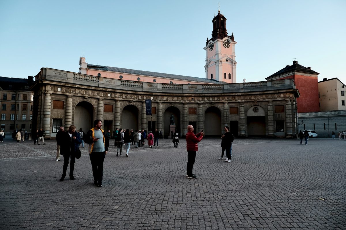 A view from Yttre Borggården in Gamla Stan, Stockholm, with people standing in the courtyard and the tower of Stockholm Cathedral visible beyond the palace buildings.